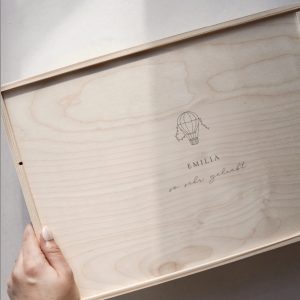Minimalistische Aufnahme einer hellen Holzbox mit Gravur ‚JULIUS‘ und Heißluftballon-Motiv von einer Hand gehalten auf einem hellen Greige-Hintergrund mit einem sanften Sonnenstrahl.