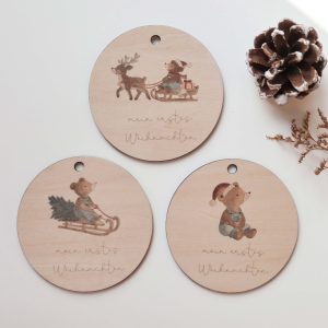Holzanhänger „Mein erstes Weihnachten“ mit drei weihnachtlichen Bärenmotiven als Aquarelldruck, dekoriert mit Tannenzapfen und Zweigen.