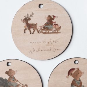 Holzanhänger „Mein erstes Weihnachten“ mit drei weihnachtlichen Bärenmotiven als Aquarelldruck, dekoriert mit Tannenzapfen und Zweigen.