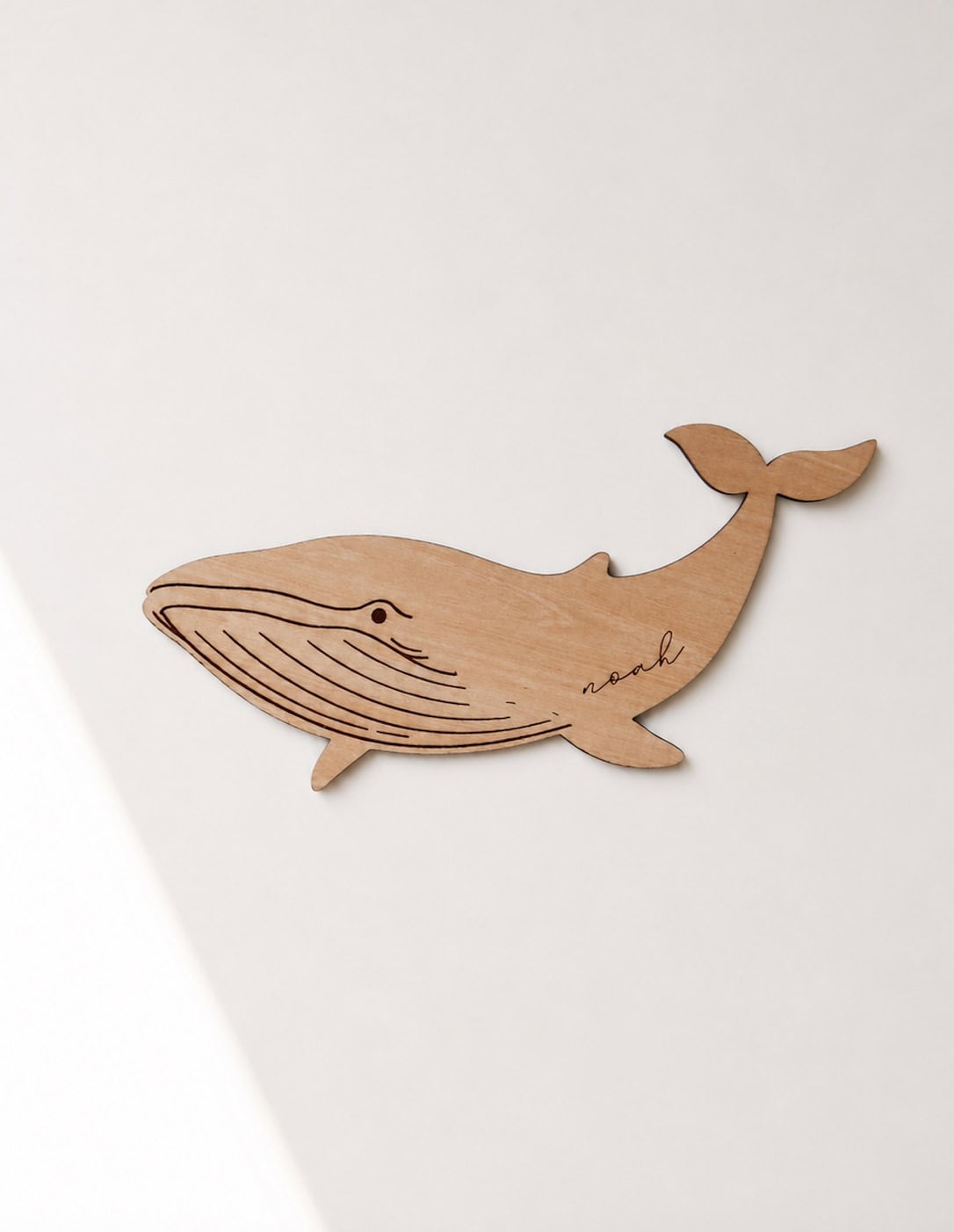 Schild aus hellem Holz in Form eines Wals mit dem Namen Noah eingraviert in minimalistischem Hintergrund