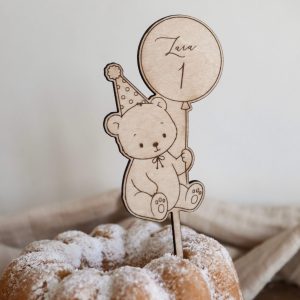 Das Bild zeigt einen Caketopper aus hellem Holz mit dem Motiv eines kindgerechten Bärs mit Partyhut und einem Ballon in der Hand, welcher den Namen "Lara" und die Zahl "1" ziert. Der Kuchenstecker steckt in einem hellen Kuchen Kuchen mit Puderzucker