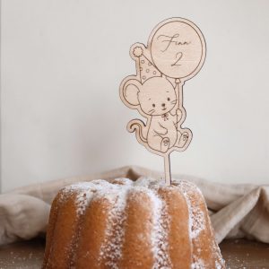 Das Bild zeigt einen Caketopper aus hellem Holz mit dem Motiv einer kindgerechten Maus mit Partyhut und einem Ballon in der Hand, welcher den Namen "Finn" und die Zahl "2" ziert. Der Kuchenstecker steckt in einem hellen Kuchen Kuchen mit Puderzucker