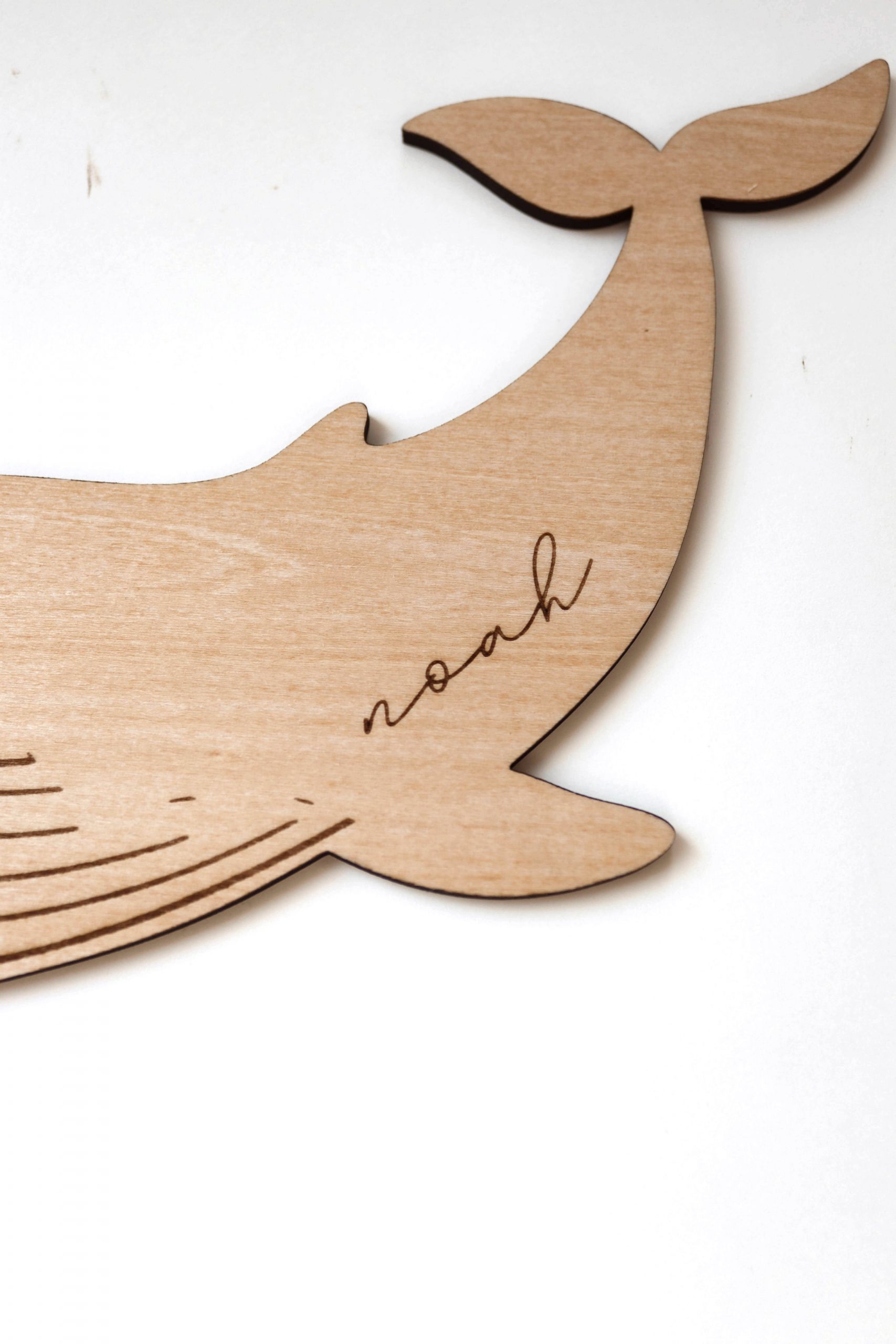 Schild aus hellem Holz in Form eines Wals mit dem Namen Noah eingraviert in minimalistischem Hintergrund
