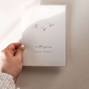 personalisierte mutterpasshülle mit schlichtem design mit drei fliegenden gänsen und dem text "mutterpass Sarah schmitt" von einer hand gehalten in minimalistischem hintergrund mit natürlichem sonnenlicht