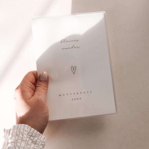 personalisierte mutterpasshülle mit schlichtem design eines herzens und dem text "kleines wunder - mutterpass anna" in minimalistischem hintergrund mit natürlichem sonnenlicht