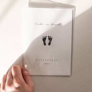 personalisierte mutterpasshülle mit schlichtem design von Babyfüßen und dem text "liebe im bauch - mutterpass anna" in minimalistischem hintergrund mit natürlichem sonnenlicht