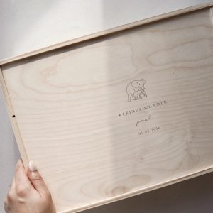 Minimalistische Aufnahme einer hellen Holzbox mit Gravur ‚Matteo‘ und Elefanten-Motiv auf einem hellen Greige-Hintergrund mit einem sanften Sonnenstrahl.