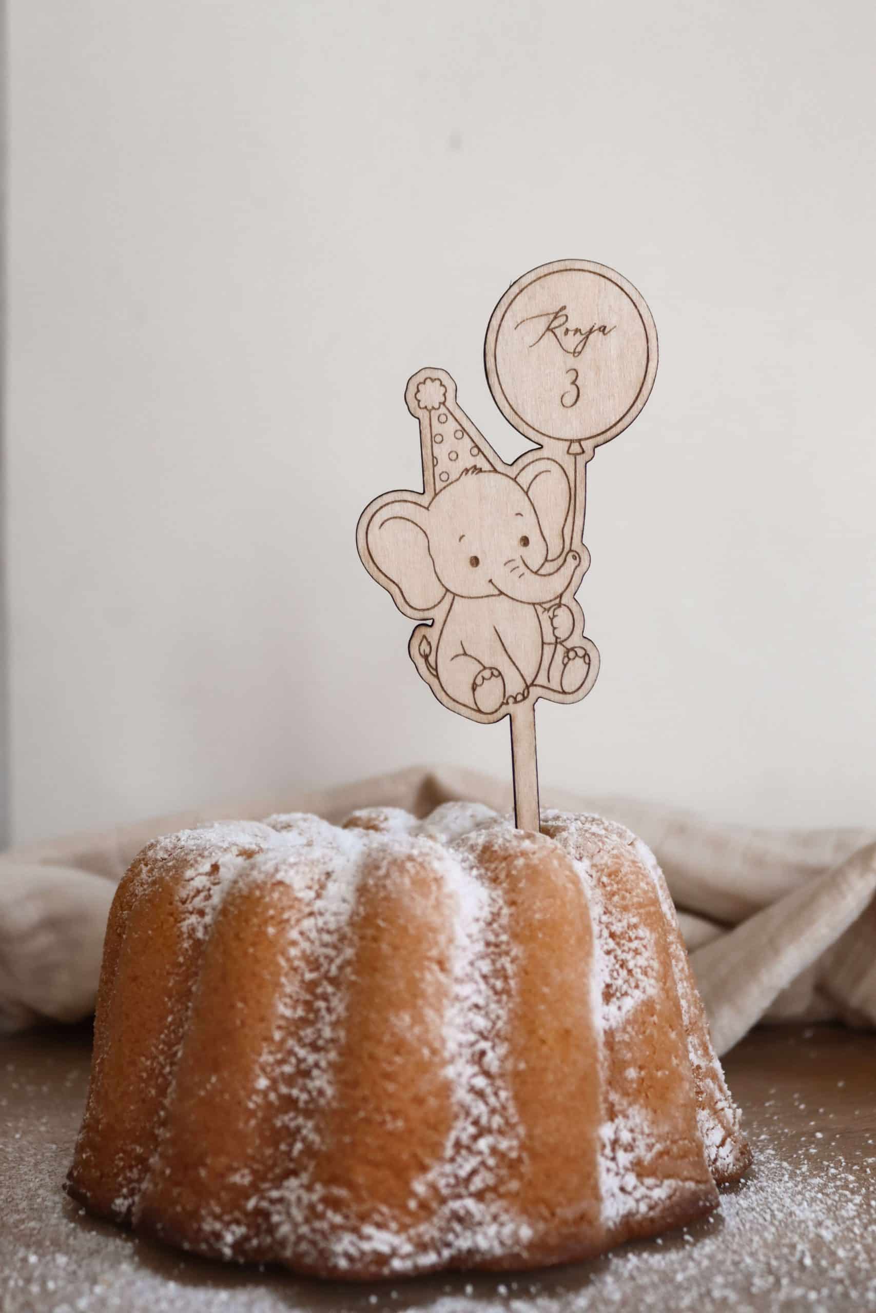Das Bild zeigt einen Caketopper aus hellem Holz mit dem Motiv eines kindgerechten Elefanten mit Partyhut und einem Ballon in der Hand, welcher den Namen "Ronja" und die Zahl "3" ziert. Der Kuchenstecker steckt in einem hellen Kuchen Kuchen mit Puderzucker